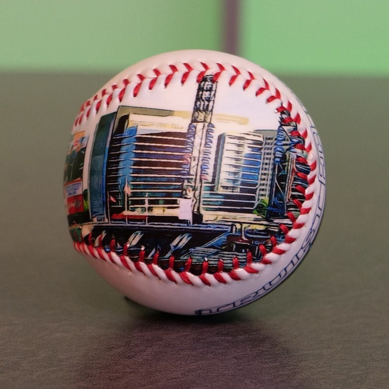 May include: White baseball with red stitching and a printed image of a city skyline. The skyline is a mix of blue, gray, and white buildings.