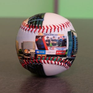 May include: A white baseball with red stitching and a printed image of a city skyline and a baseball team. The team's logo is a red "A" with a white outline. The team's name is "Atlanta Braves" and the team's sponsor is "Northside Hospital".