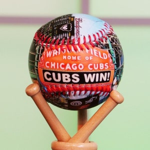 May include: A baseball with a colorful design featuring the Chicago Cubs logo and the words "Wrigley Field, Home of Chicago Cubs, Cubs Win!" The baseball is on a wooden stand.