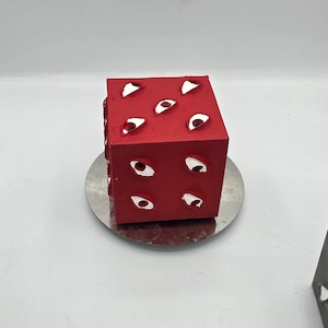 May include: A red cube with white eyes cut out of each side. The cube is sitting on a silver circular plate.