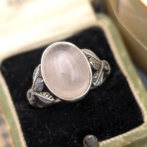 Könnte beinhalten: Ein silberner Ring mit einem ovalen, hellrosa Edelstein. Der Ring hat ein Blattmuster auf dem Band und wird in einer grünen Vintage-Schmuckschatulle mit schwarzem Samtinterieur präsentiert.