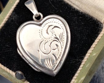May include: A silver heart-shaped locket featuring an engraved swirling floral pattern on the front surface. The locket includes a small silver bail at the top for attachment and is presented inside a small, open, light green vintage jewelry box.