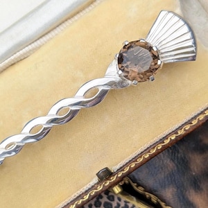 May include: A silver kilt pin with a twisted design and a brown gemstone. The pin is in a vintage box with a yellow interior and a patterned exterior. The pin is a decorative accessory.