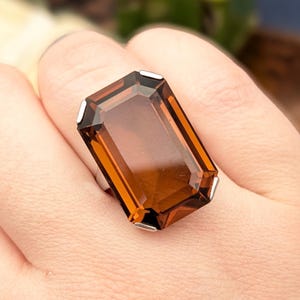 May include: A large rectangular faceted amber-coloured gemstone ring set in a simple silver-toned metal prong setting. The stone features a deep cognac hue with clear transparency and sharp, clean edges, showcasing a classic emerald-cut style design.