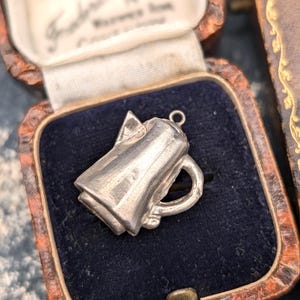 Könnte beinhalten: Ein silberfarbener Metallanhänger in Form einer kleinen Kaffeekanne oder eines Kruges. Der Anhänger verfügt über einen Henkel an einer Seite, einen Ausguss auf der gegenüberliegenden Seite und eine kleine runde Öse oben zur Befestigung.