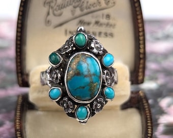 May include: A silver ring featuring a large oval turquoise center stone with brown matrix, surrounded by six smaller round turquoise stones and silver floral accents. The ring is displayed inside a vintage brown velvet-lined watch box.