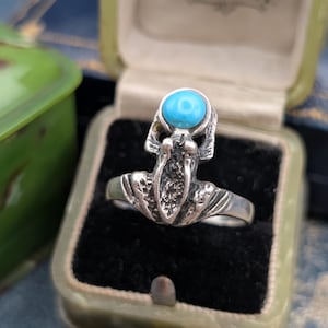 May include: A silver ring featuring a textured, stylised frog design with a small, round, bright blue turquoise stone set at the top. The ring is displayed inside a vintage, light green, rectangular jewellery box with black velvet lining.