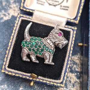 May include: A silver-toned brooch shaped like a terrier dog, featuring a body covered in small green circular stones and a single red circular stone for the eye. The brooch is displayed inside a dark blue hinged jewelry box with gold trim.