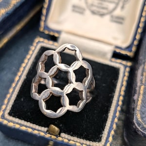 Op de afbeelding: Een zilveren ring met een opengewerkt ontwerp bestaande uit onderling verbonden cirkelvormige en zeshoekige vormen. Het metaal heeft een gepolijste afwerking en de structuur creëert een herhalend geometrisch patroon op de ring.