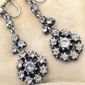 Può includere: Un paio di orecchini pendenti antichi color argento. Ogni orecchino presenta un gruppo di gemme chiare e scintillanti che circondano una pietra centrale più grande. Gli orecchini hanno una chiusura a vite e sono esposti in un portagioie color crema.