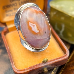 Op de afbeelding: Een zilveren ring met een grote, ovaalvormige steen. De steen heeft een mix van kleuren, waaronder oranje, wit en paars. De ring is gezet in een zilveren band en wordt gepresenteerd in een vintage sieradendoosje.