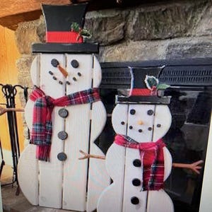 May include: Two wooden snowman decorations, one large and one small, standing in front of a stone fireplace. The snowmen are painted white with black buttons and a red and black plaid scarf.