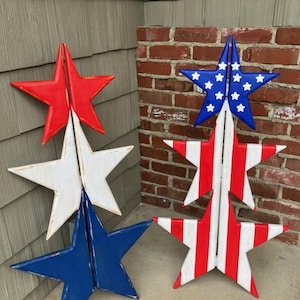 May include: Three sets of wooden stars in red, white, and blue, stacked on top of each other. The stars are painted in a distressed style, giving them a vintage look.