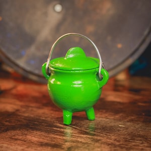 May include: A small, bright green cauldron with a matching lid and a silver handle. The cauldron has three short legs and is sitting on a wooden surface. The background is blurred, with a dark, metallic object visible.