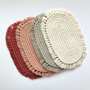 May include: Oval macrame placemats in shades of red, pink, gray, and cream. Each placemat features a woven design with a fringed edge. The placemats are arranged in a fan shape, showcasing the different colors and textures.