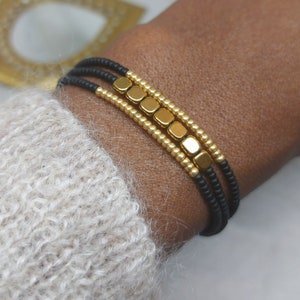 May include: A layered bracelet with black elastic cord and gold square beads.