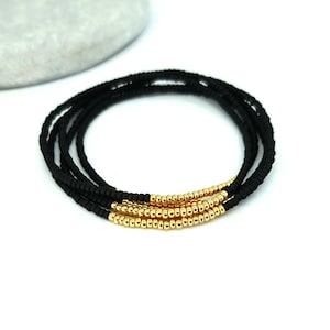 May include: A multi-strand bracelet composed of small, matte black beads. Each of the three strands features a segment of metallic gold-toned beads arranged in a row, creating a contrasting colour pattern against the black bead base.