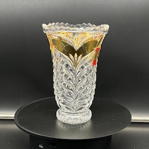 May include: A clear glass vase with a gold rim and intricate cut glass design. The vase is on a black rotating display stand.