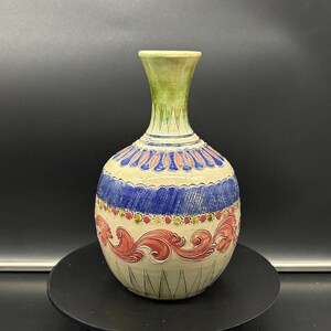 Może przedstawiać: Ceramiczny wazon o okrągłym korpusie i wąskiej szyjce. Wazon jest ozdobiony kolorowym wzorem w kolorze niebieskim, czerwonym i złotym. Wazon stoi na czarnym cokole.