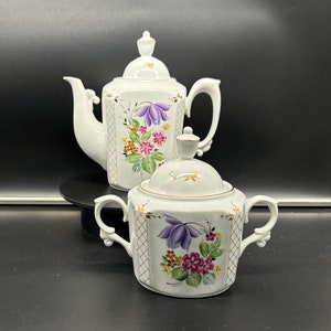 Pode incluir: Um conjunto de bule e açucareiro de porcelana branca com um design floral. O bule tem um corpo quadrado com uma borda dourada e o açucareiro tem um corpo redondo com uma tampa dourada.