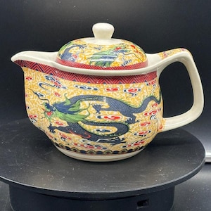 Op de afbeelding: Een witte theepot van keramiek met een drakenontwerp. De theepot heeft een gele achtergrond met rode, blauwe en groene accenten. De draak is afgebeeld in een vliegende pose met zijn vleugels uitgespreid. De theepot heeft een deksel met een knop bovenop.