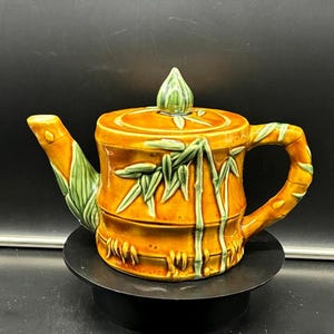 May include: A ceramic teapot with a bamboo design. The teapot is primarily orange with green bamboo accents. The handle, spout, and lid are also orange with green detailing. The teapot is sitting on a black stand.
