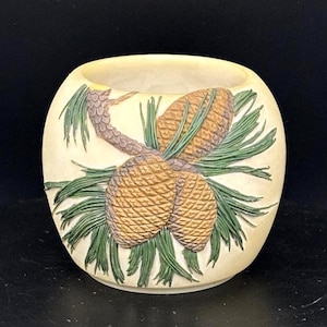 May include: Cream-colored, round decorative bowl with a detailed relief of pine cones and needles. The pine cones are brown, and the needles are green. The bowl has a smooth, unglazed finish, and the design is raised.