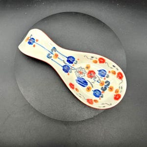 May include: A ceramic spoon rest with a white base, red trim, and a floral design in blue, orange, and red. The spoon rest is oval-shaped and designed to hold a spoon while cooking.