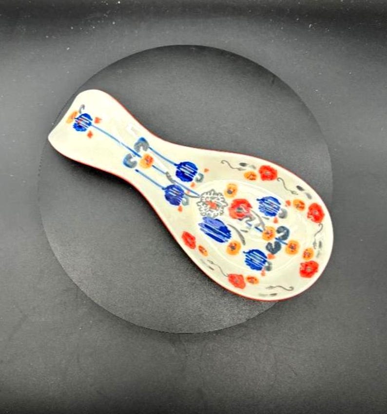 May include: A ceramic spoon rest with a white base, red trim, and a floral design in blue, orange, and red. The spoon rest is oval-shaped and designed to hold a spoon while cooking.