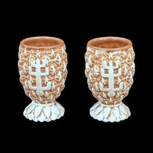 May include: Two ceramic goblets featuring a textured, light brown pineapple-like exterior with a raised white emblem on the front. Each goblet has a smooth, light brown interior rim and a wide, scalloped white base that supports the cup.