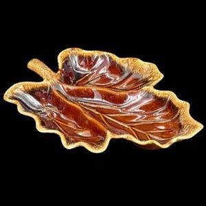 May include: A ceramic leaf-shaped serving dish with a glossy finish. The dish is primarily brown with a gold-coloured trim around the edges. The leaf's veins are subtly visible, adding detail to the design. The dish is set against a black background.