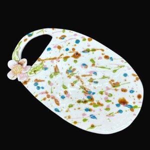 Puede incluir: Una bandeja de servicio ovalada de cerámica blanca con asa, que presenta un patrón de salpicaduras de pintura multicolor en tonos azul, verde, naranja y rosa, acentuado por una flor esculpida rosa y amarilla unida al área del asa.
