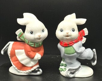 May include: A pair of white porcelain bunny figurines ice skating. The female bunny is wearing a red dress with a green and white scarf. The male bunny is wearing a blue suit with a red scarf. Both bunnies have black skates.