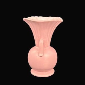 May include: A light pink ceramic vase with a bulbous base and a flared, ruffled top. The vase has a handle and vertical ribbed detailing. The vase is set against a black background.