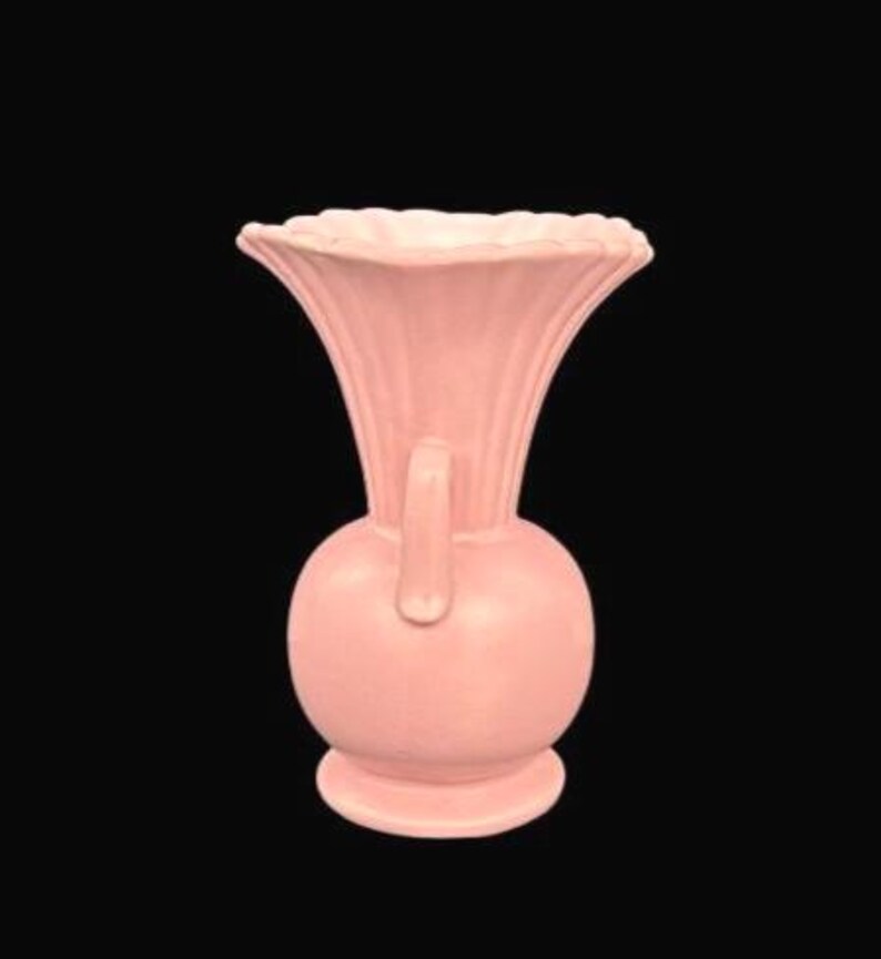 May include: A light pink ceramic vase with a bulbous base and a flared, ruffled top. The vase has a handle and vertical ribbed detailing. The vase is set against a black background.