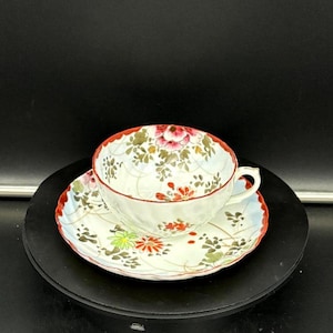 May include: A white teacup and saucer set with a floral design. The cup and saucer have a red trim and are decorated with pink, red, and green flowers and leaves. The set is placed on a black surface.