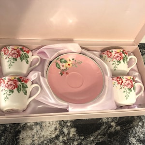 May include: A boxed tea set featuring four white teacups and a pink saucer, each adorned with a floral pattern of red and pink roses and green leaves. The set is presented in a pink box with a satin lining.