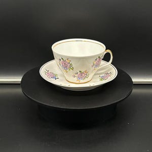 May include: A white teacup and saucer with a floral pattern in pink, blue, and yellow. The cup and saucer have a gold trim.