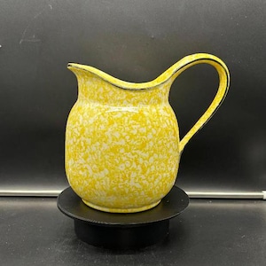 Puede incluir: Una jarra de cerámica amarilla con un patrón moteado blanco y un borde negro. La jarra tiene un asa y está sentada sobre un pedestal negro.