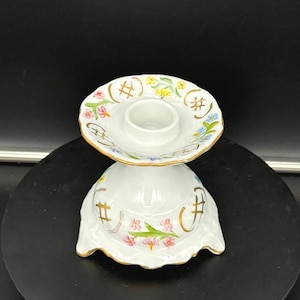 Puede incluir: Candelabro de cerámica blanca con un diseño floral en rosa, amarillo y azul. El candelabro tiene un borde festoneado y ribete dorado. El diseño incluye el símbolo de la almohadilla.