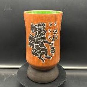 May include: A brown ceramic cup with a green rim and a black and white design of a figure holding a staff. The figure is surrounded by a circle of eyes.