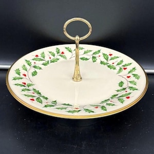 Può includere: Un vassoio da portata a due livelli con manico e bordo dorati. I piatti bianchi sporchi sono decorati con foglie di agrifoglio verdi e bacche rosse. Il vassoio è progettato per servire dessert o antipasti.