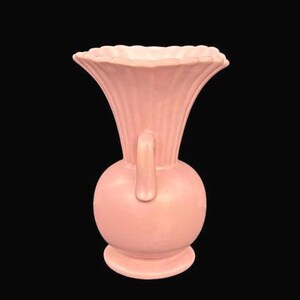 May include: A vintage pink vase with a bulbous base, a handle, and a flared, scalloped rim. The vase has vertical ribbed detailing on the upper portion. The vase is made of ceramic or porcelain and is set against a black background.