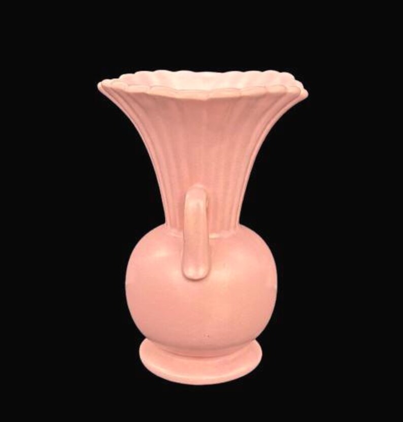 May include: A vintage pink vase with a bulbous base, a handle, and a flared, scalloped rim. The vase has vertical ribbed detailing on the upper portion. The vase is made of ceramic or porcelain and is set against a black background.