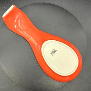 May include: A bright orange ceramic spoon rest with a white oval center. The spoon rest has a long handle and a curved shape, designed to hold a spoon while cooking. The spoon rest is made of ceramic.