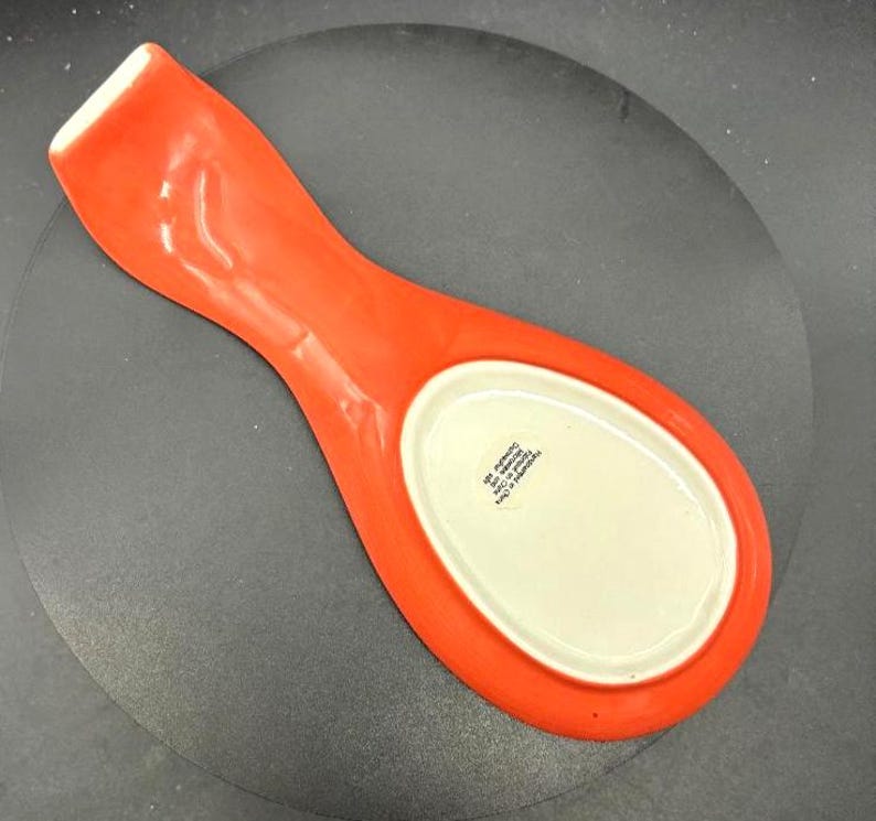 May include: A bright orange ceramic spoon rest with a white oval center. The spoon rest has a long handle and a curved shape, designed to hold a spoon while cooking. The spoon rest is made of ceramic.