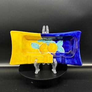 Op de afbeelding: Een gele en blauwe asbak van keramiek met een geschilderd ontwerp van citroenen en bladeren.