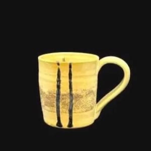 May include: Yellow ceramic mug with a curved handle. The mug has a textured surface with two vertical black stripes and a patterned design. The mug is set against a black background.