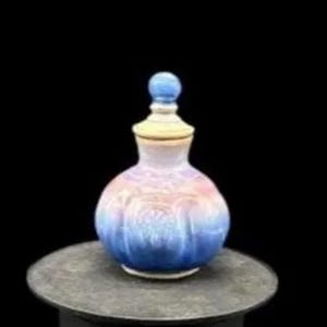 May include: A decorative, hand-painted glass bottle with a lid. The bottle features a gradient of colors, transitioning from blue at the base to pink and yellow at the top. The lid is topped with a blue sphere. The bottle is displayed on a dark gray surface.