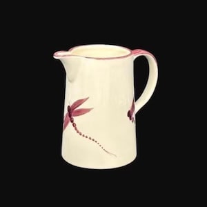 May include: Cream-colored ceramic pitcher with a curved handle and spout. The pitcher is decorated with maroon dragonfly designs. The rim and handle are trimmed with maroon. The pitcher is set against a black background.